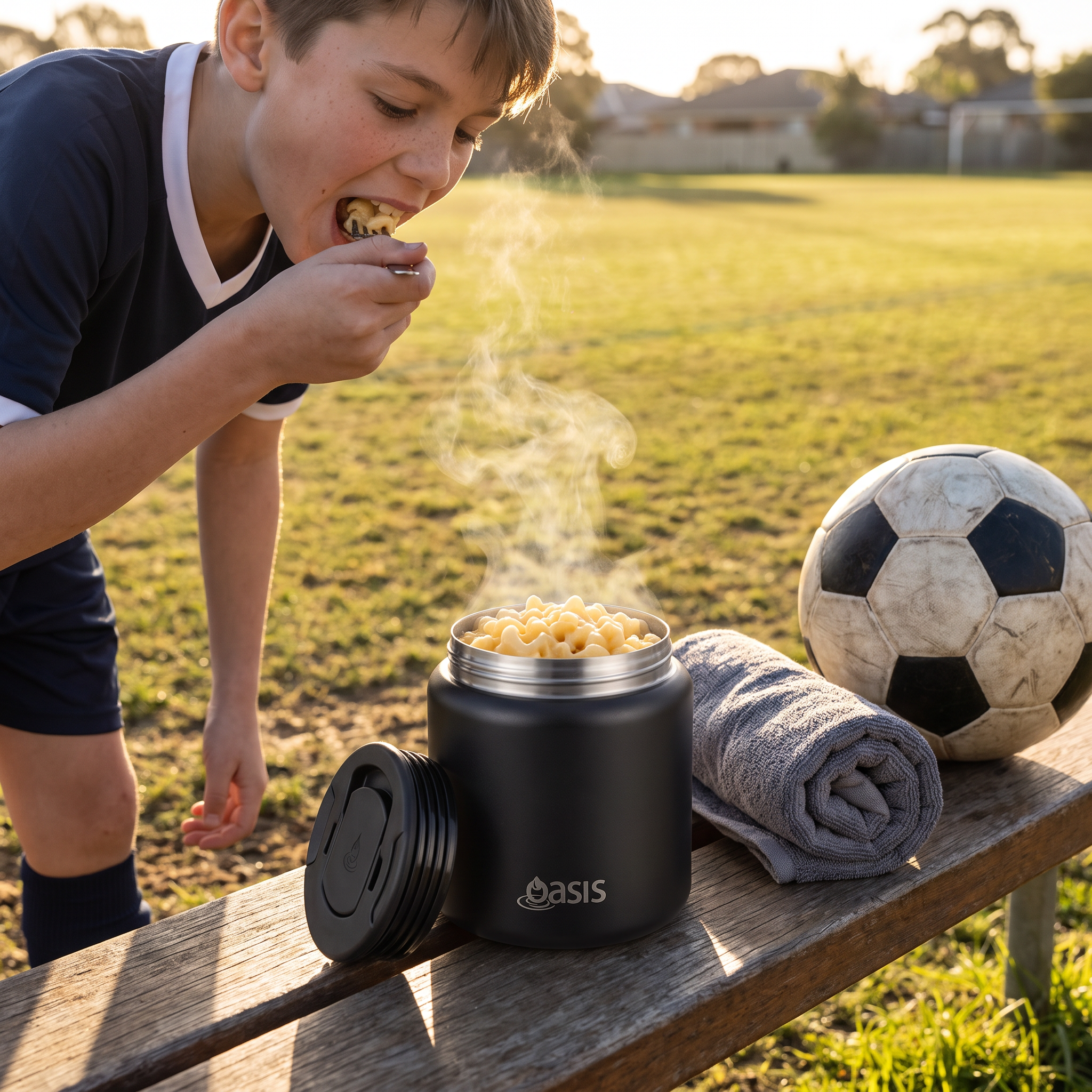 A boy in a soccer jersey eats from a black Oasis insulated snack container on a wooden bench at a soccer field. A soccer ball and a rolled towel are beside the container, with steam rising from the food.