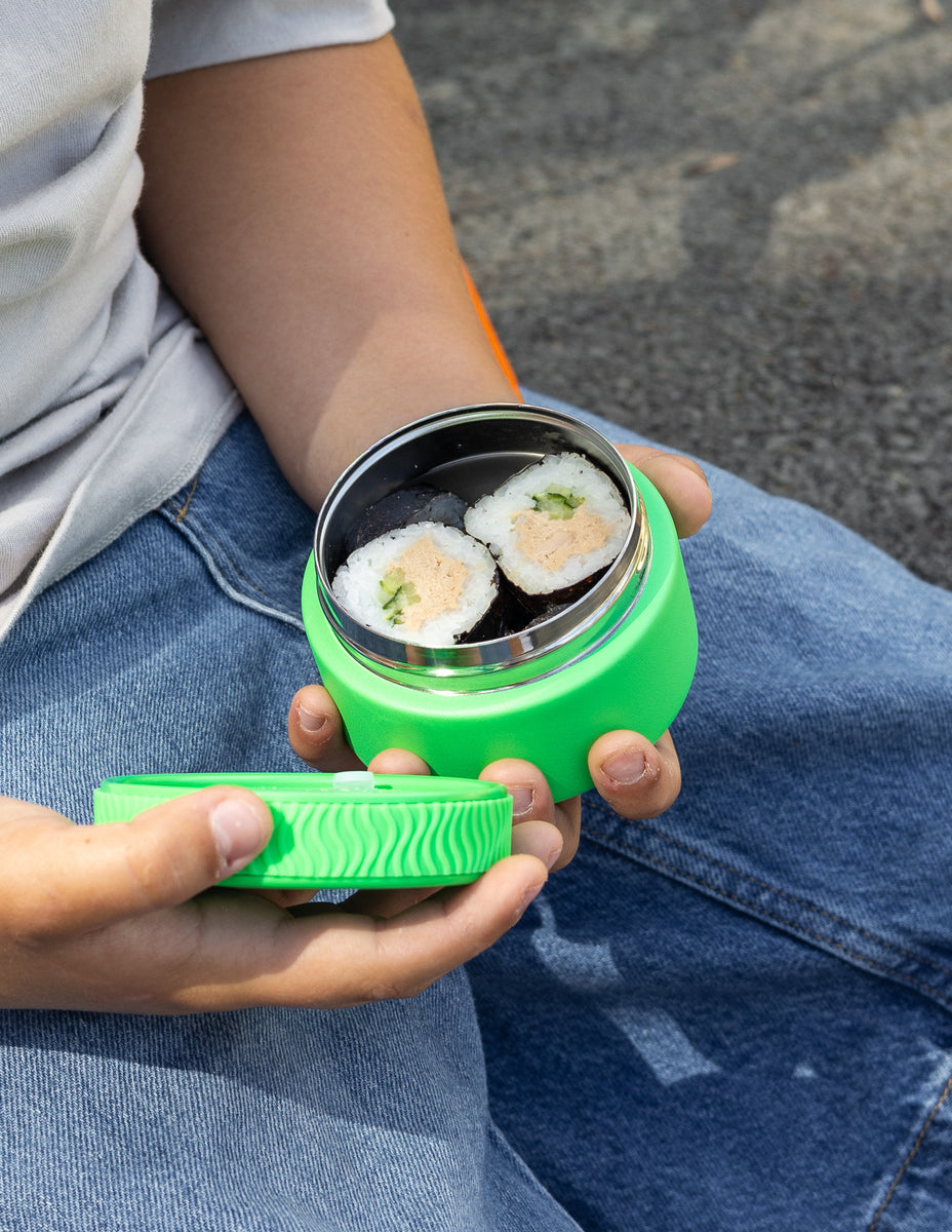 Person holding a green container with sushi inside, sitting on a concrete surface.