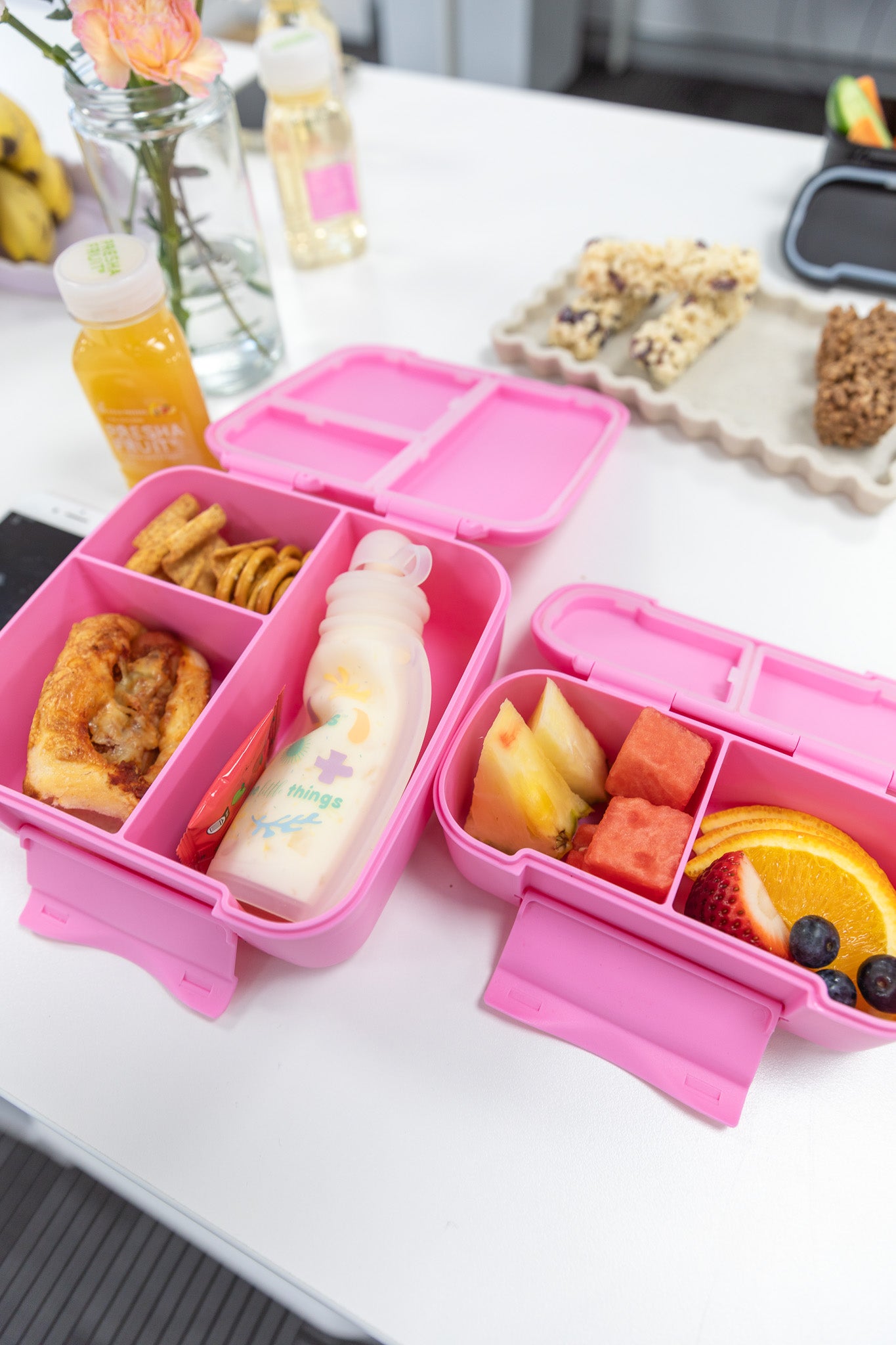 Pink bento box with compartments containing snacks and a bottle on a white table.