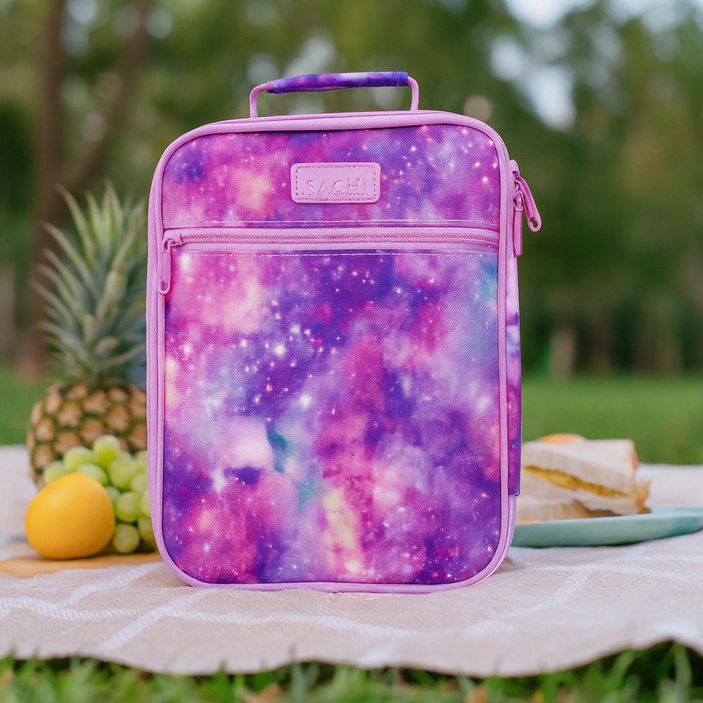 Colorful galaxy-patterned lunch bag on a picnic blanket with fruits and sandwiches.