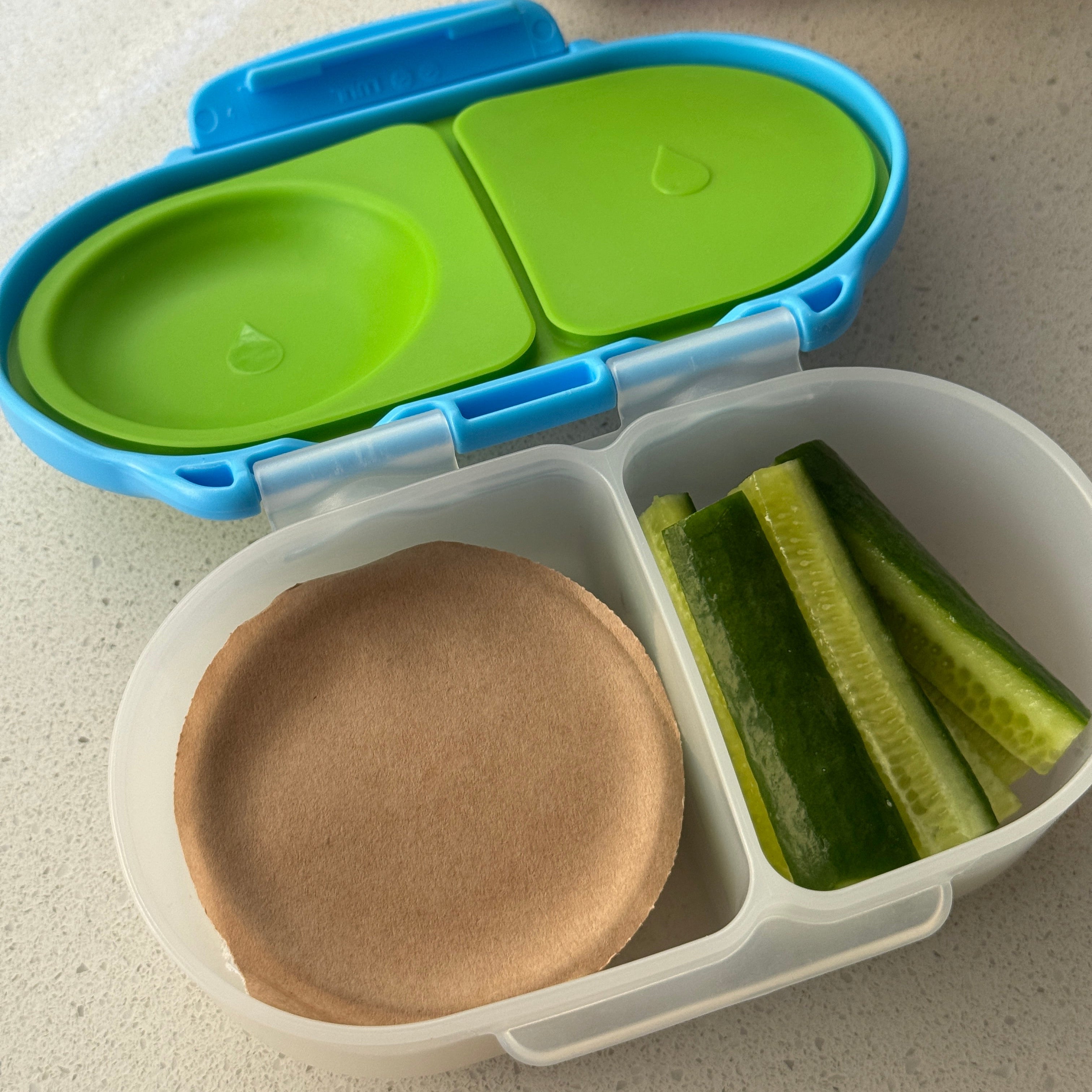 A close-up of a compartment bento lunch box with a translucent white container, a bright blue lid, and green insert plates. The box is open and contains cucumber slices in one compartment and a brown paper plate in another.