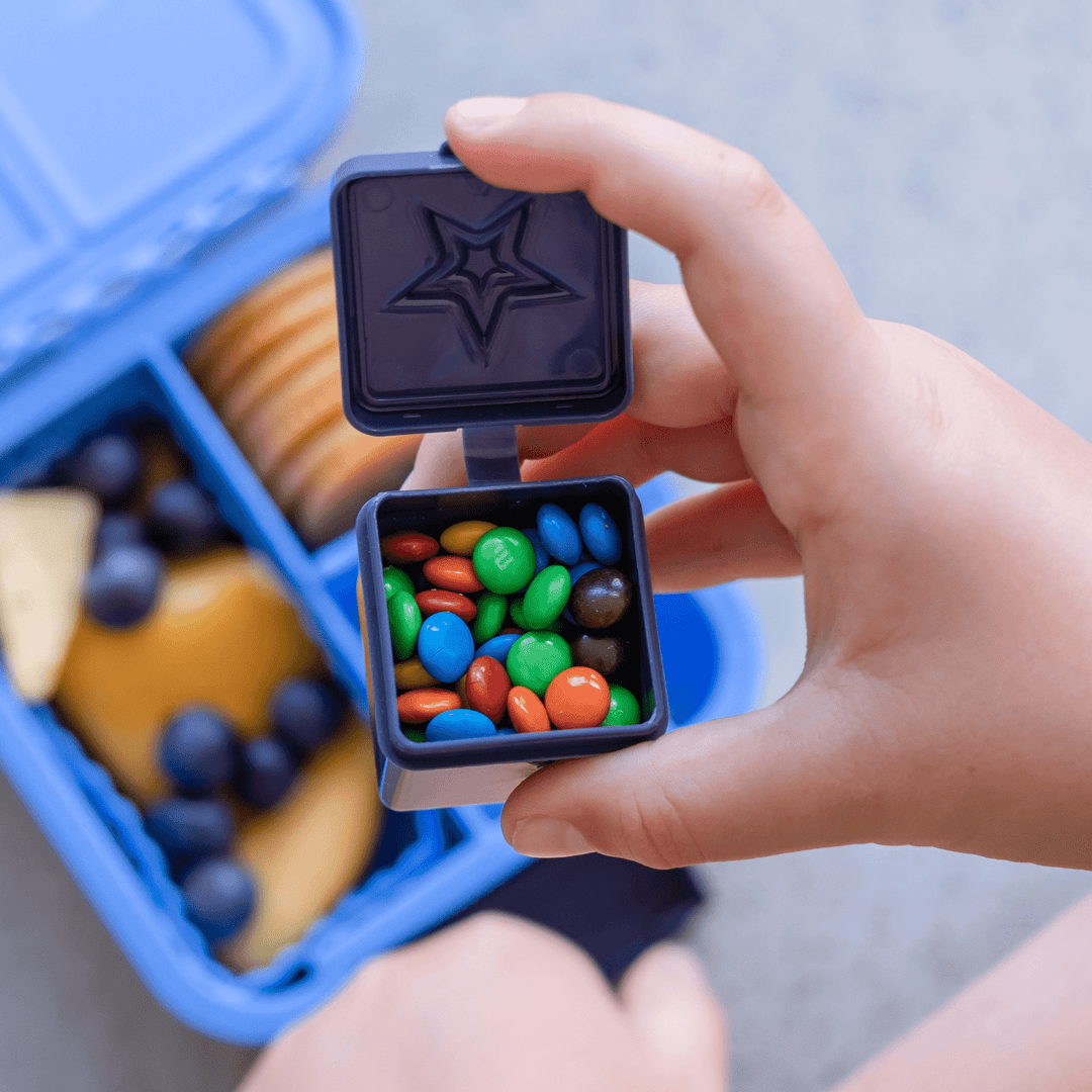 Hand holding a small black box with colorful candies, with a blue lunchbox in the background.