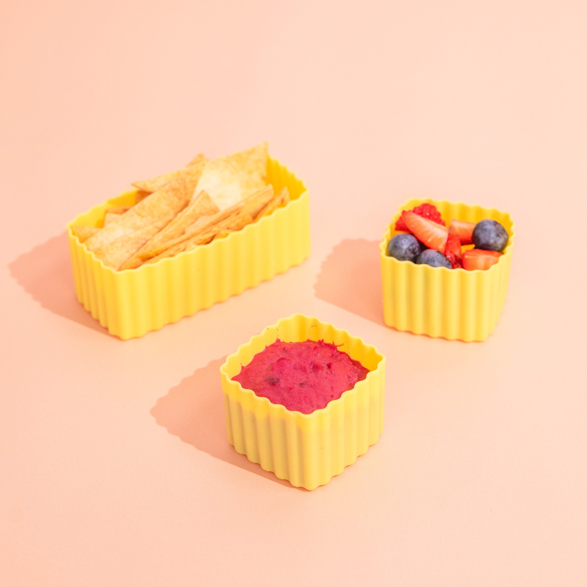 Three yellow silicone food containers filled with different foods on a pink background