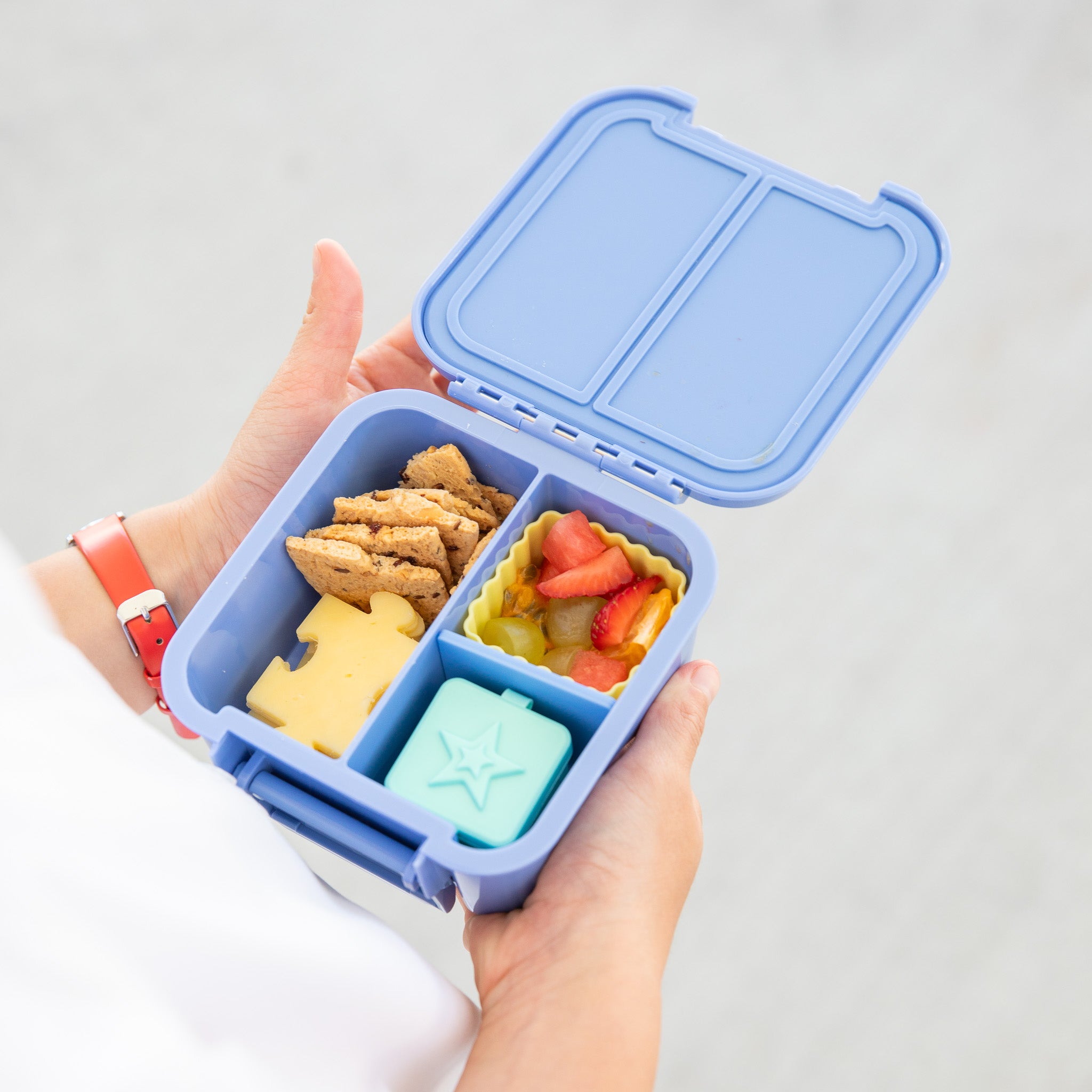 Hand holding a blue bento box with snacks on a light gray background