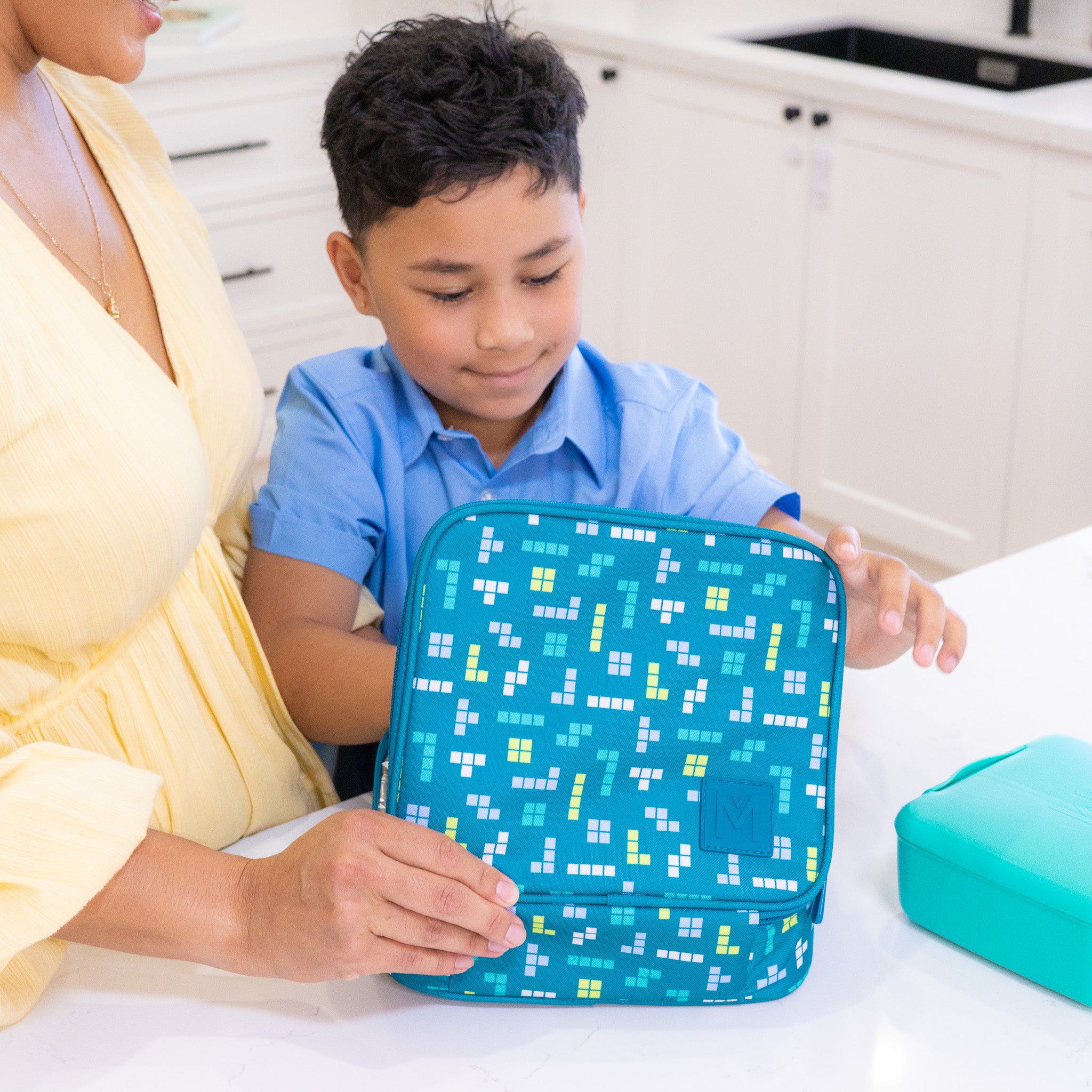 Child holding a blue lunch bag with pixelated design, sitting next to an adult in a kitchen.