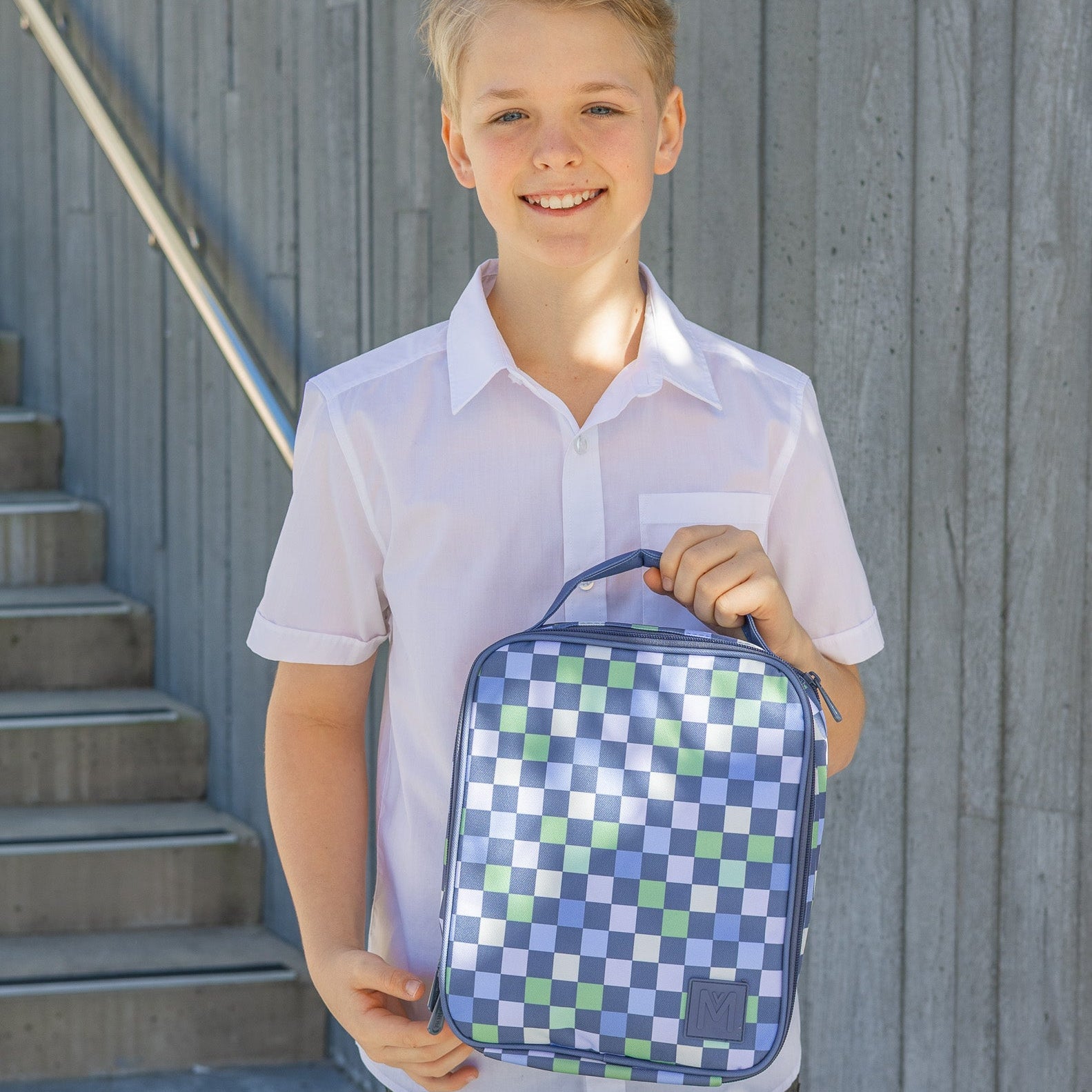 Young boy holding a checkered lunch bag against a gray wall.