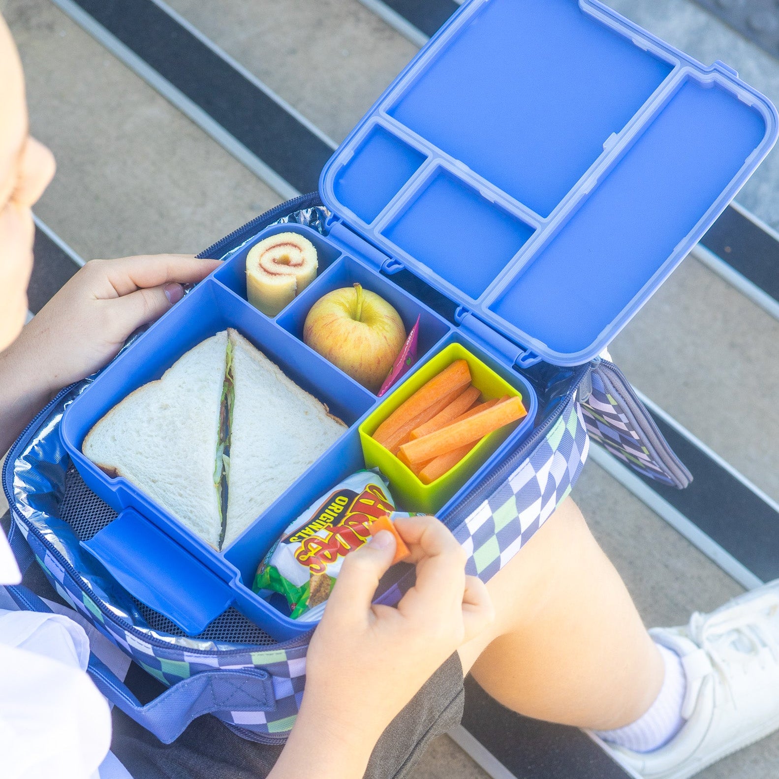 Blue lunch box with compartments containing a sandwich, apple, carrots, and other snacks.