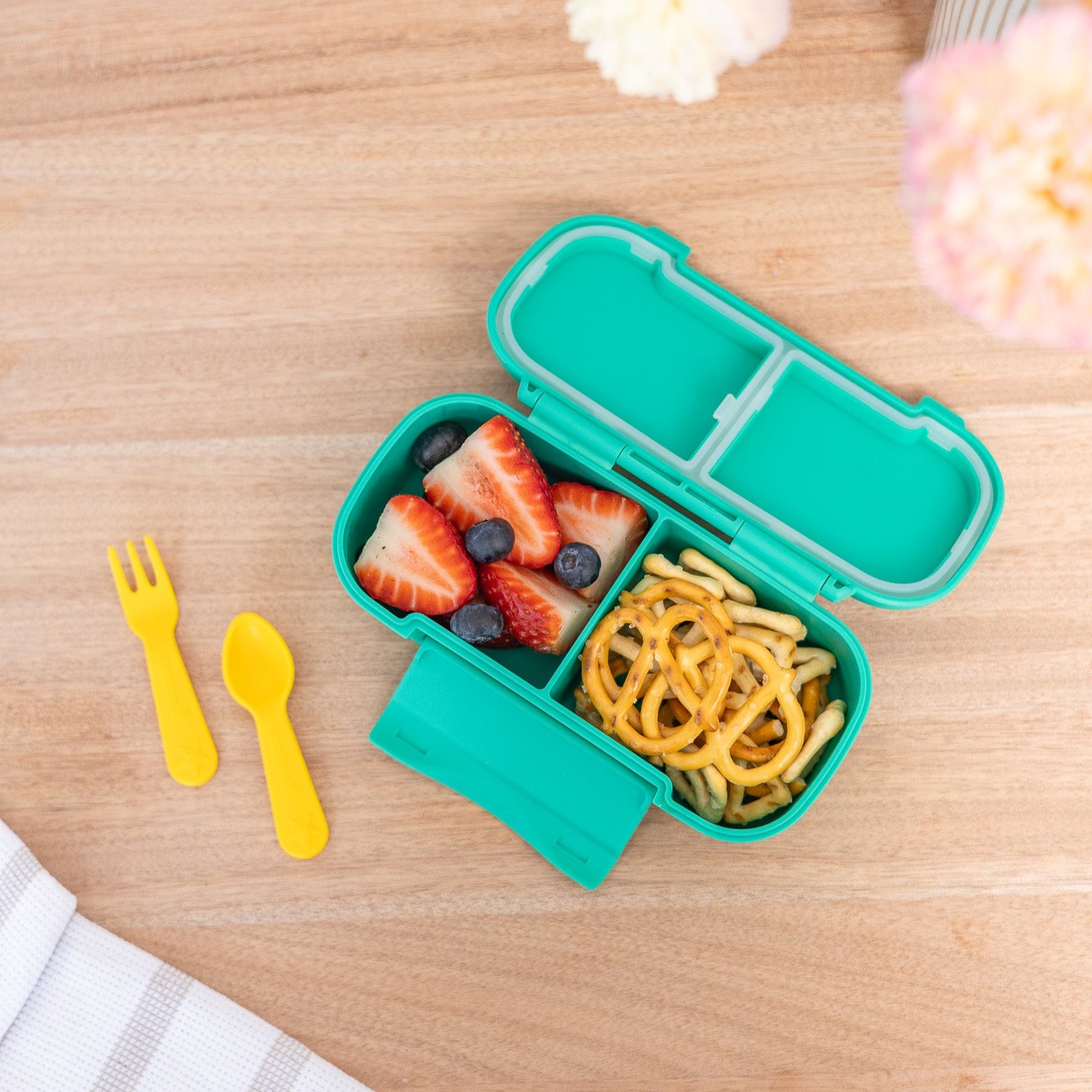 Green bento box with food compartments on a wooden table with yellow fork and spoon.