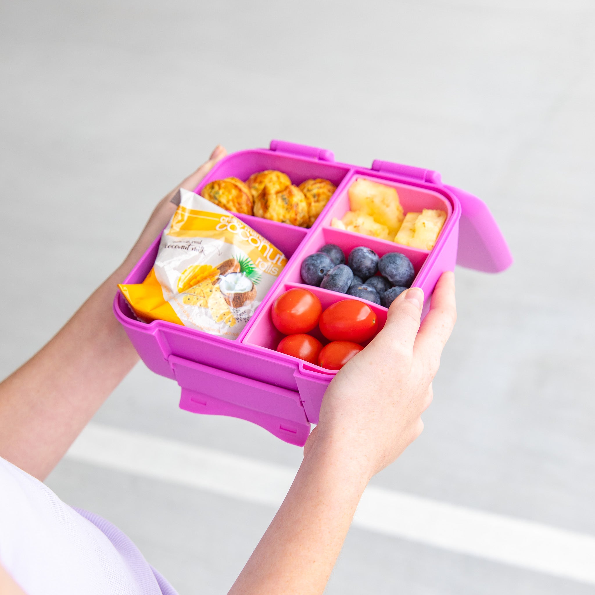 Person holding a pink bento box with compartments filled with snacks and fruits on a light gray background