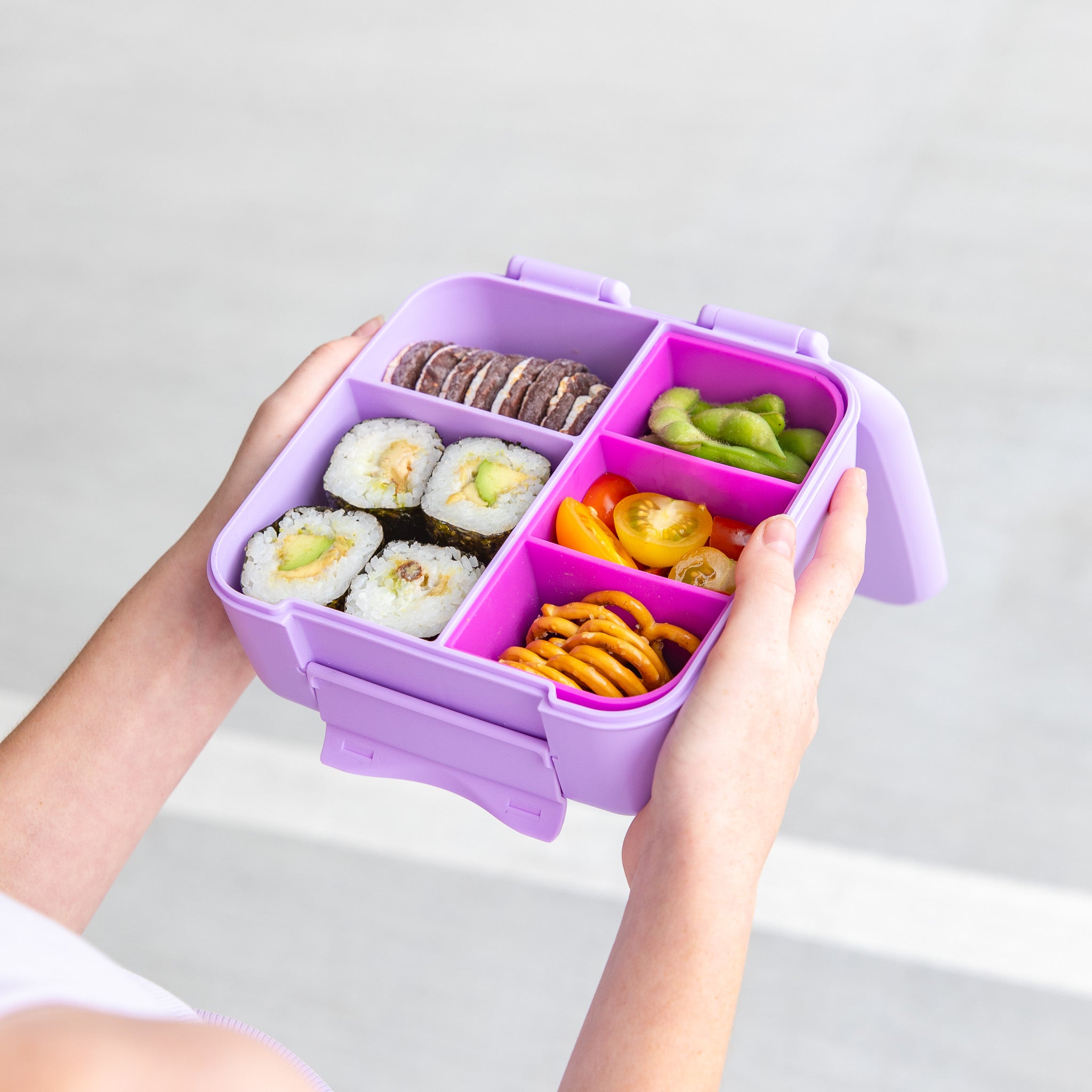 Purple bento box with compartments filled with various snacks held by a person against a light gray background