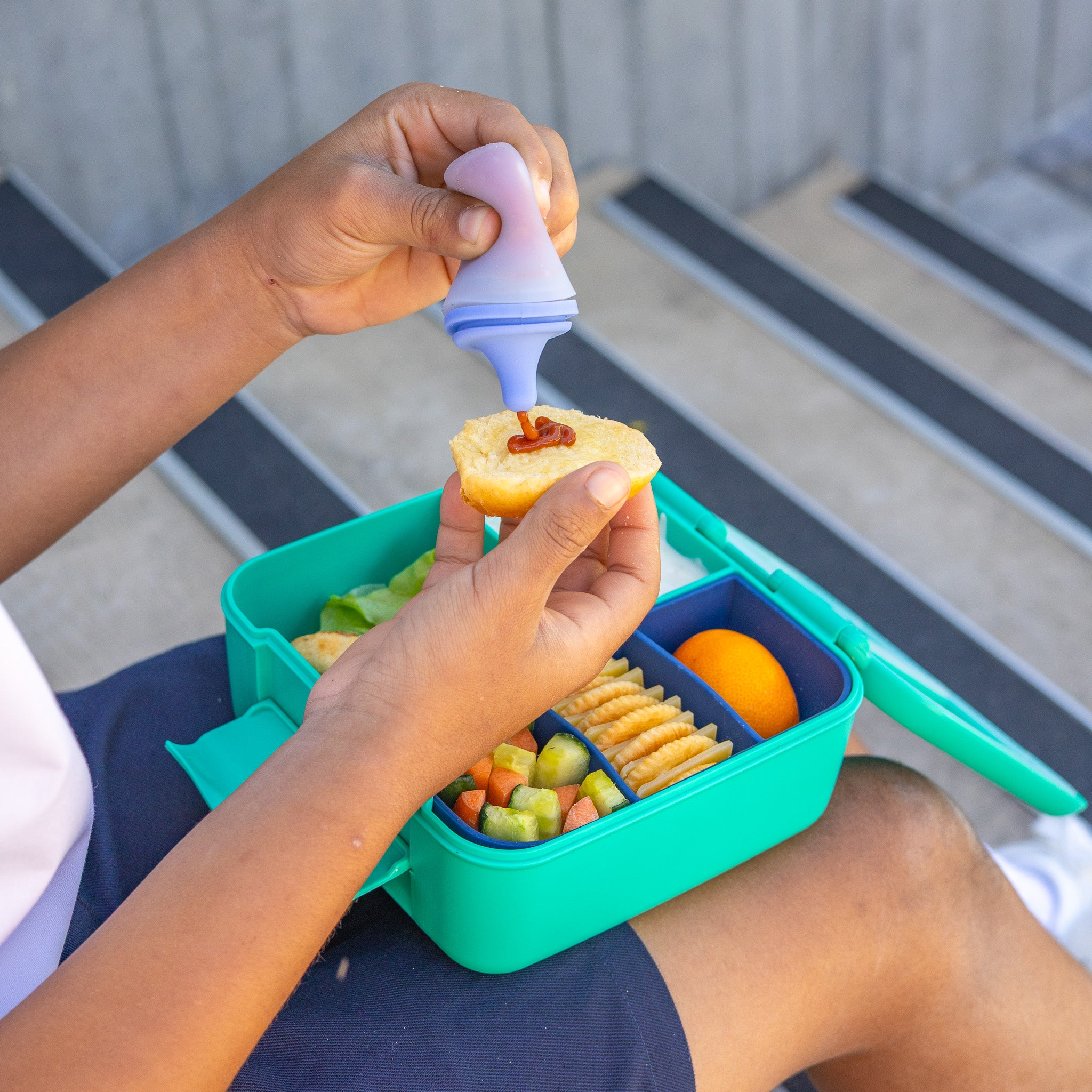 Person adding a snack to a colorful bento box on a bench.