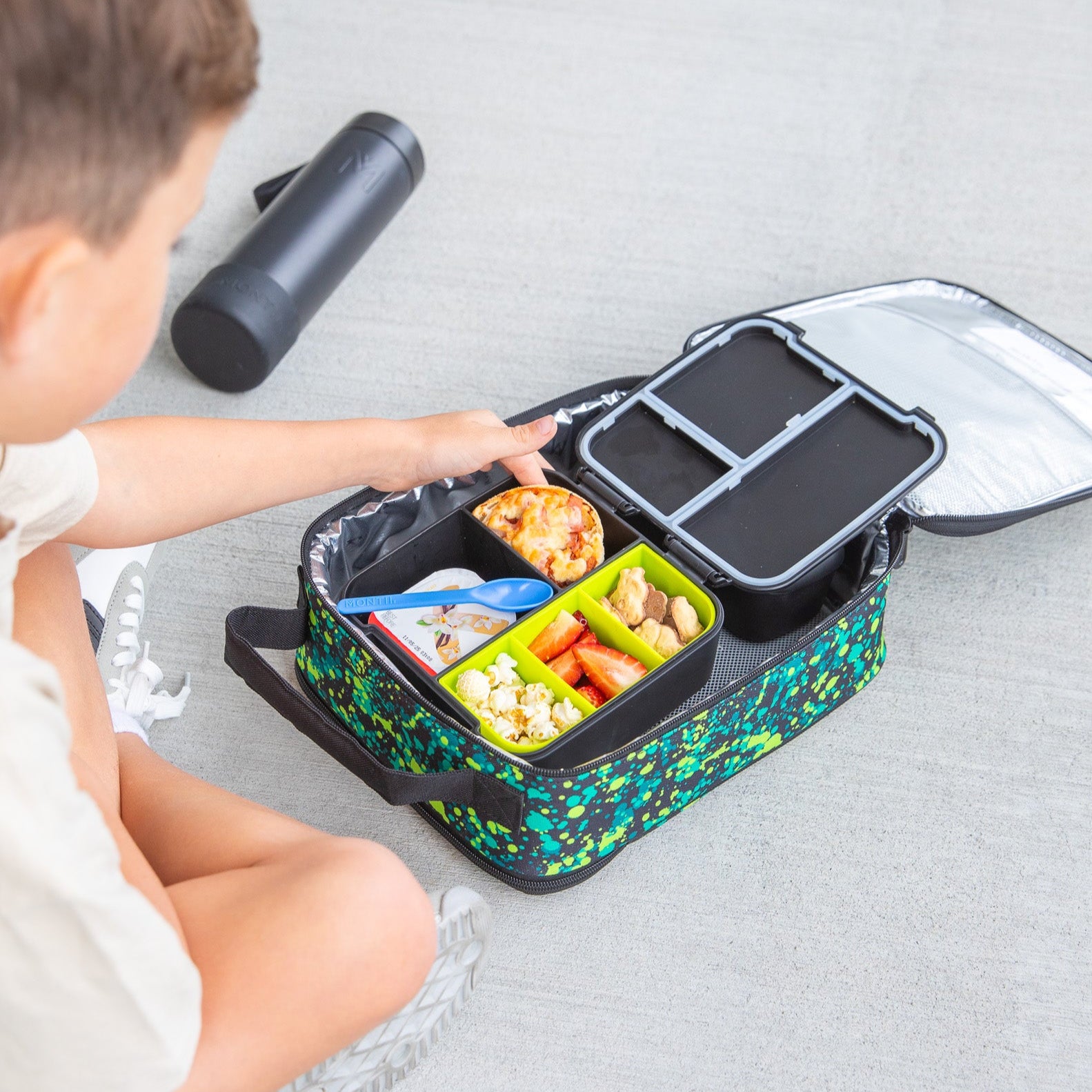 Child opening a colorful bento box with food on a light gray surface
