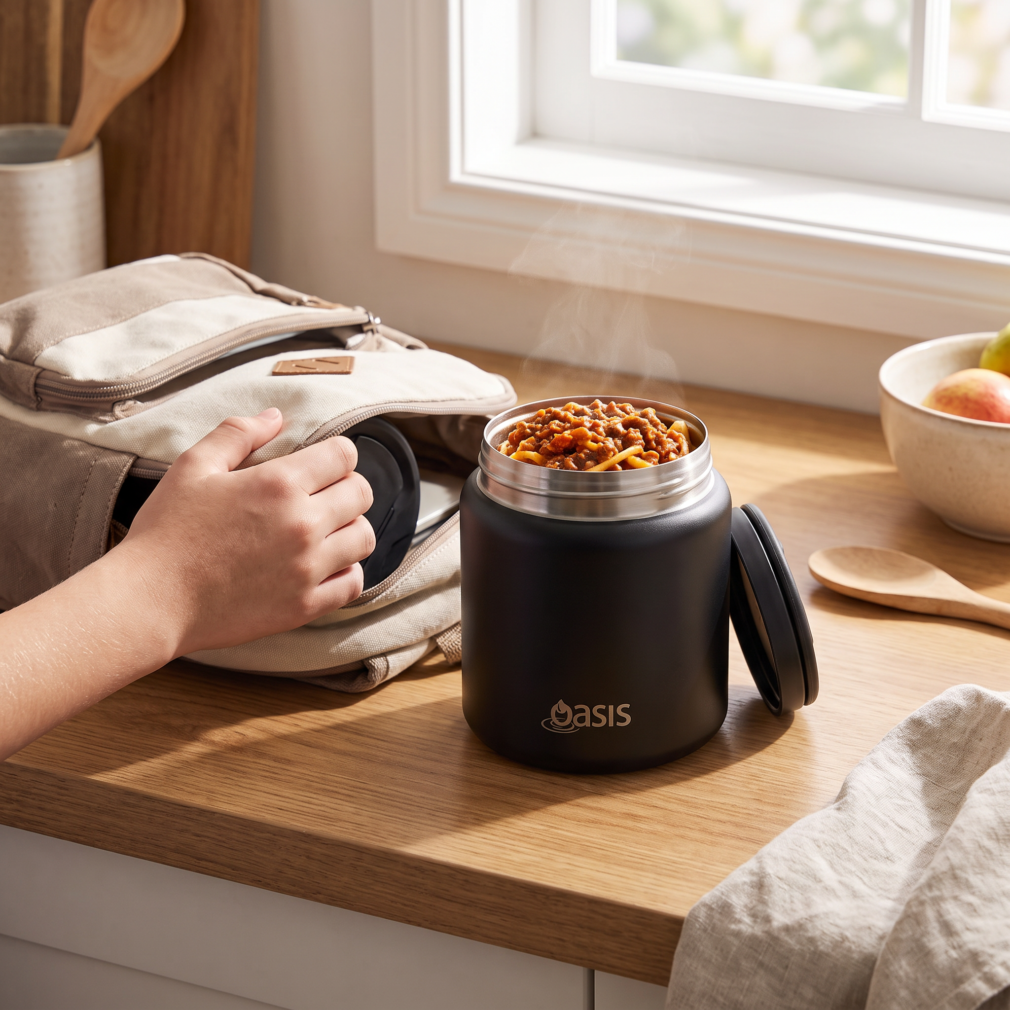 A black Oasis insulated food jar filled with food sits on a wooden countertop next to a beige backpack, with a hand reaching toward it in a bright kitchen setting.
