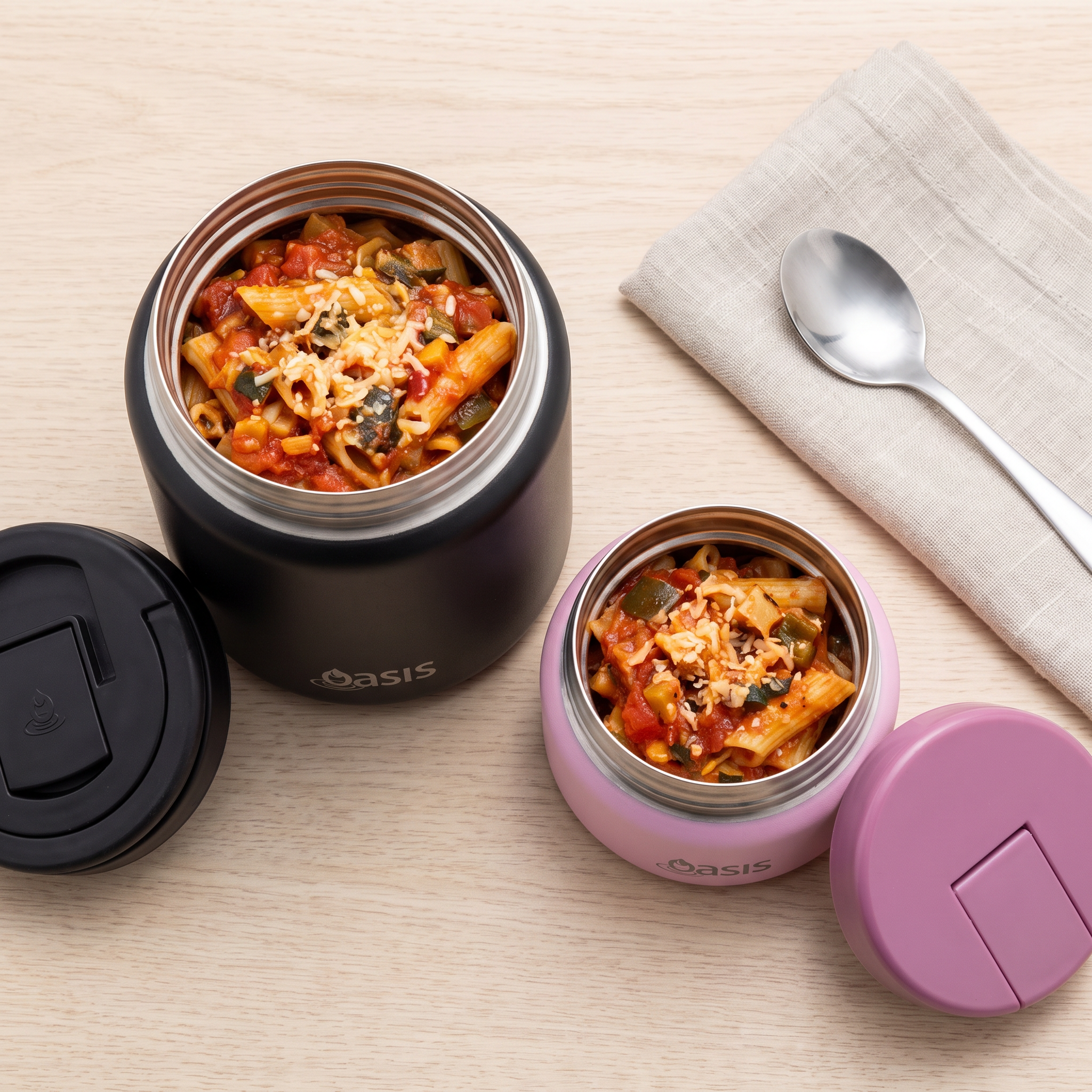 Two insulated food containers on a light wood surface, one black and one pink, both open with pasta and sauce inside. A black lid, a spoon, and a beige cloth napkin are arranged around them.