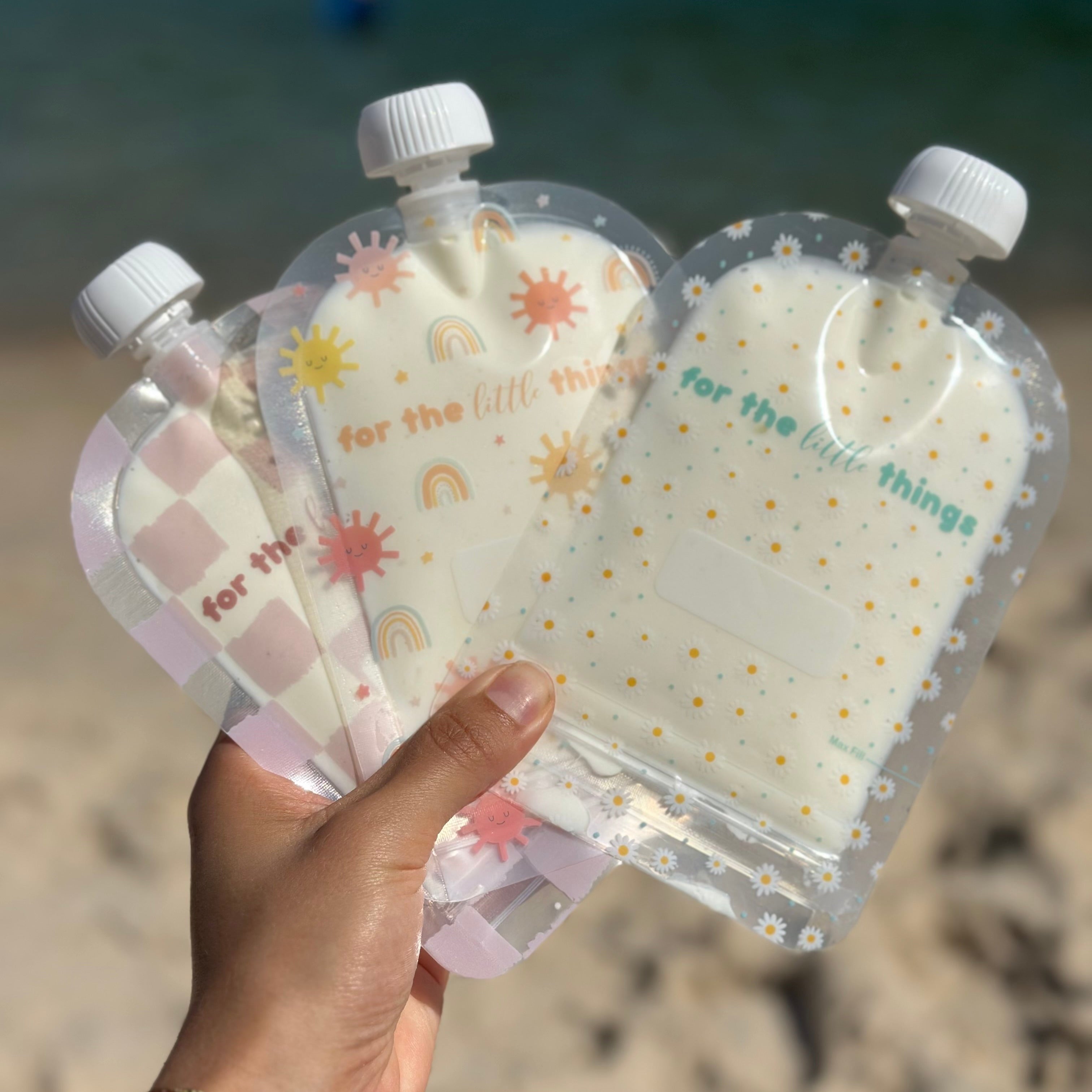 Three clear plastic pouches with colorful designs held by a hand on a beach.