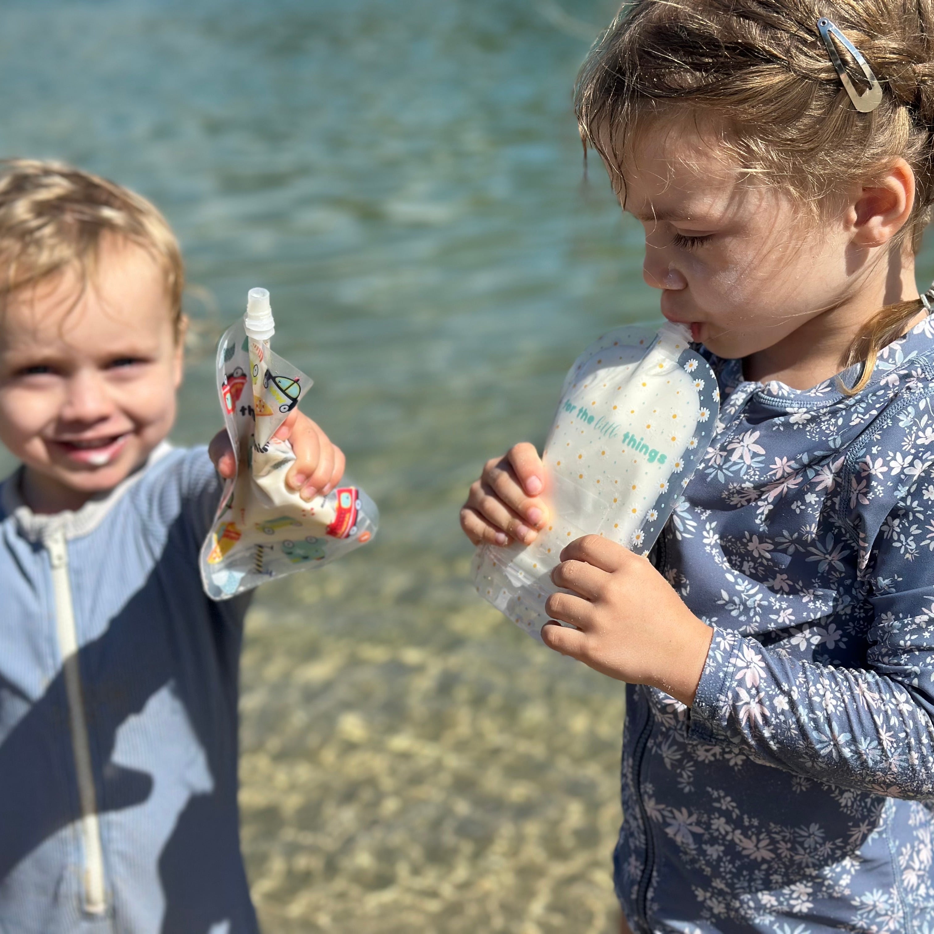 Two children by a body of water, holding plastic bottles with trash inside.