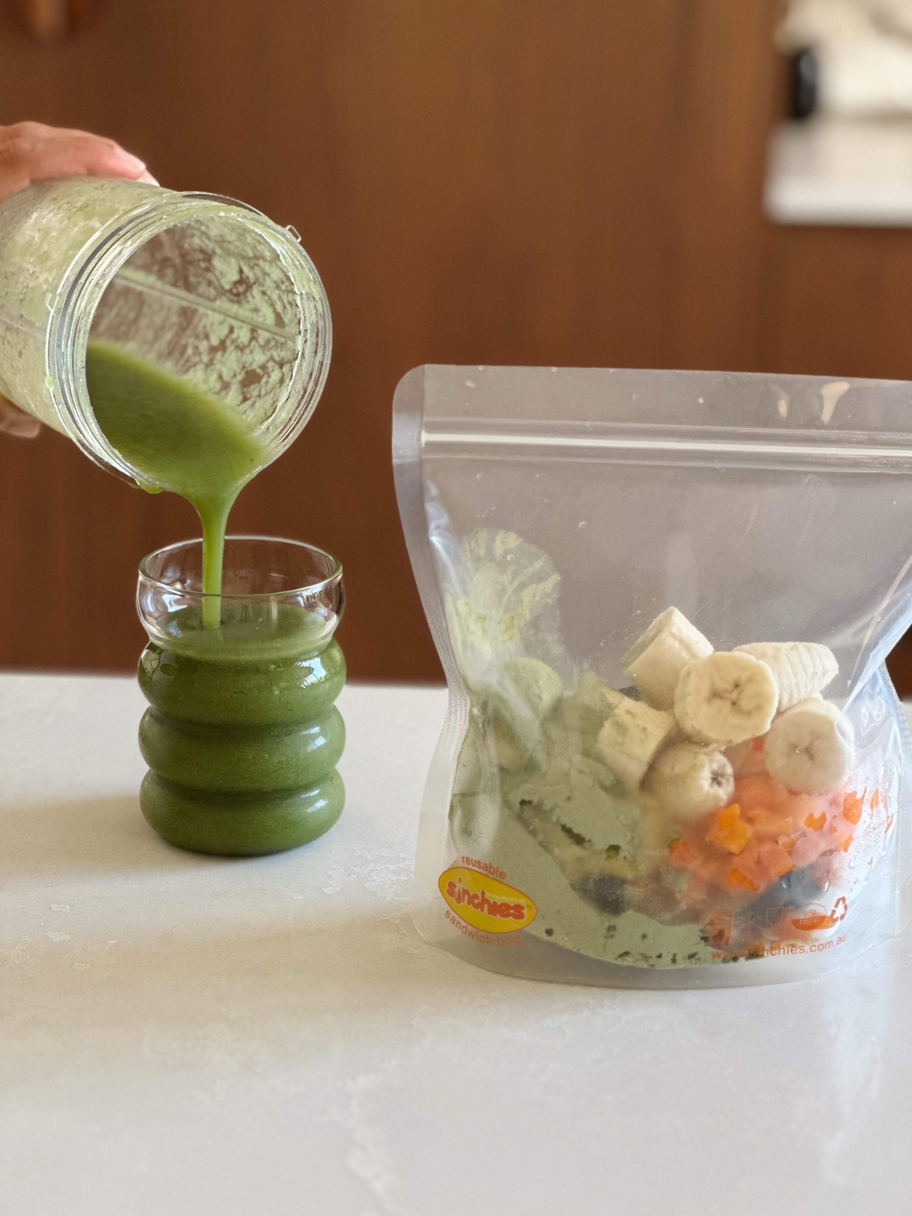Green smoothie being poured into a glass with a package of ingredients on a white surface.